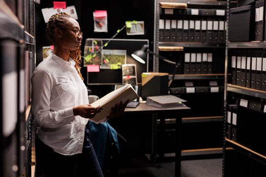 Law enforcement professional reading investigation materials in detective office room. African american woman investigator holding crime case file and looking at folders in shelf