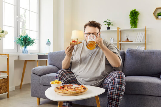 Pizza and beer choice of chubby hungry man at home, diet off. Funny overweight guy with glasses and pajamas sitting on sofa to enjoy fast food and lager in mug, takeaway cozy meals for weekend