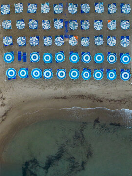 Aerial view of rows of blue and white striped and light blue beach umbrellas and sunbeds on the sandy shore of Cefalu, Sicily, Italy.