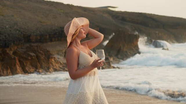 Elegant woman wearing sunhat and summer dress drinking wine by ocean waves at rocky beach enjoying holiday