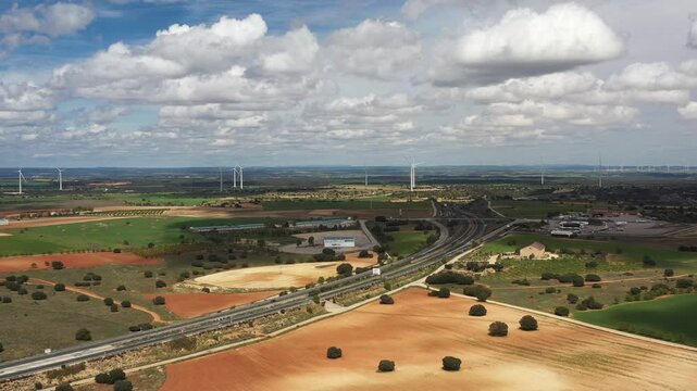 Aerial drone shot of highway intersection with vehicles in motion, surrounded by wind turbines and farmland under cloudy skies, showcasing clean energy, transportation and rural landscape harmony