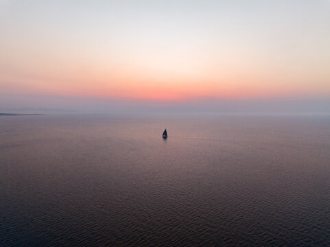 Aerial view of a lone sailboat sailing across the calm Mediterranean Sea under a soft sunset sky off the coast of Isola delle Femmine, Sicily, Italy.