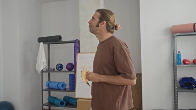 Man in brown shirt holds peeled banana with hand on hip in yoga studio, looking upward; post workout calm.