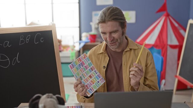 Man teacher points pencil at colorful alphabet card beside chalkboard in preschool building; early literacy warmth.