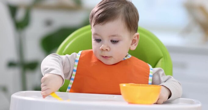 Cute little boy with spoon and bowl of baby food on feeding chair at home. Camera moving forward