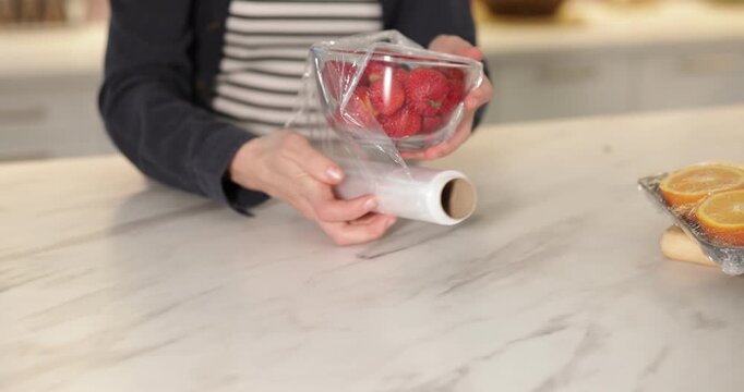 Woman wrapping bowl with fresh strawberries in cling film at light marble table, closeup