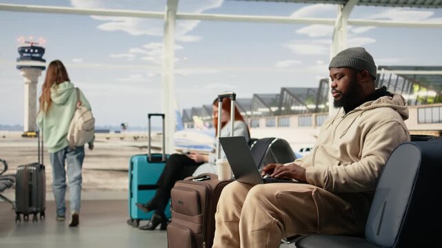 Digital nomad man, working online with laptop, in international airport terminal, seated near the boarding gate with luggage at his side. Location independent remote worker with nomadic lifestyle.