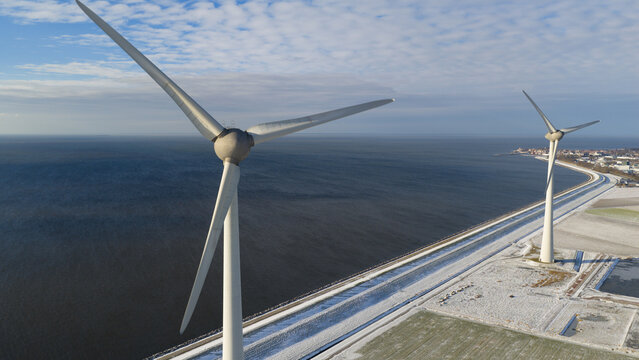 Aerial view of wind turbines at Westermeerdijk standing along the snow-covered coastline and blue water under a cloudy sky in Espel, Flevoland, Netherlands.