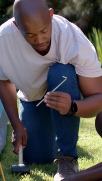 Vertical video: Placing peg kneeling dad in white T and son hitting stake securing tent with mallet