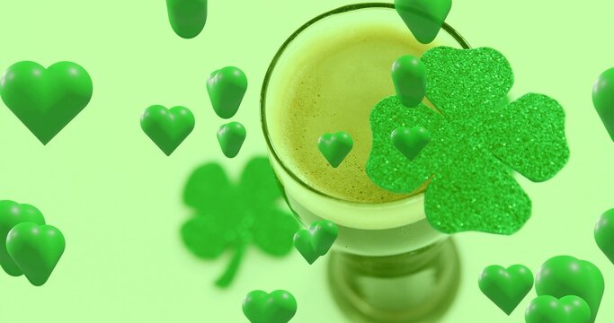 Drifting glossy green hearts circling stemmed glass holding frothy drink in studio, glitter clover