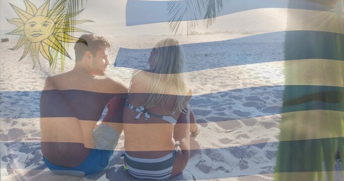 Sitting couple wearing swimwear facing ocean on sand, with blue-white stripes, yellow sun emblem