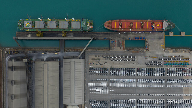 Aerial view of cargo ships docked at the industrial port terminal with rows of new cars parked in the logistics area Laem Chabang, Chon Buri, Thailand.