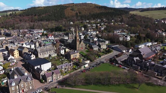 Aerial panoramic sweep over the traditional stone houses and rooftops of Peebles, capturing a prominent historic church tower and the scenic urban landscape of the Scottish Borders