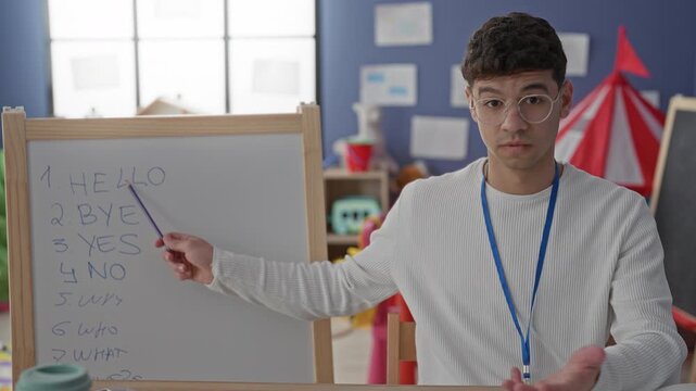 Man points pointer to whiteboard in a classroom, gesturing with open palm and wearing glasses and lanyard while teaching vocabulary; calm focus.