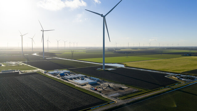 Aerial view of wind turbines standing in flat agricultural fields near Olsterdwarspad with a canal and utility station under a bright sky in Dronten, Flevoland, Netherlands.