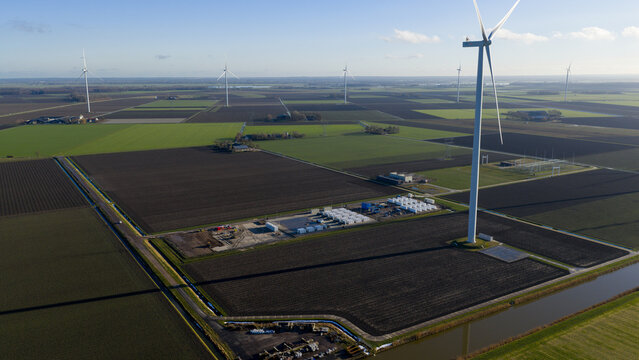 Aerial view of wind turbines standing over agricultural fields and a battery storage facility under a clear sky at Olsterdwarspad in Dronten, Flevoland, Netherlands.