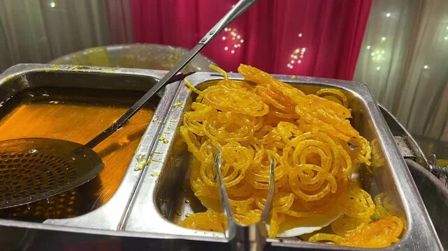 Fresh, crispy Jalebis are being deep-fried and soaked in sugar syrup at a live dessert counter during a grand festive celebration in India.