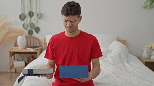 Man holding blue gift box on bed inside a building, grimacing while lifting the lid with his hands; confusion reaction.