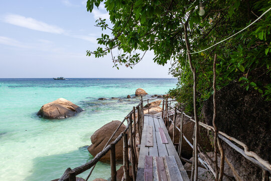 Wooden pathwalk leading to tropical beach in Koh Lipe. Turquoise waters and large rocks dot the coastline. Lush greenery frames the scenic seascape.
