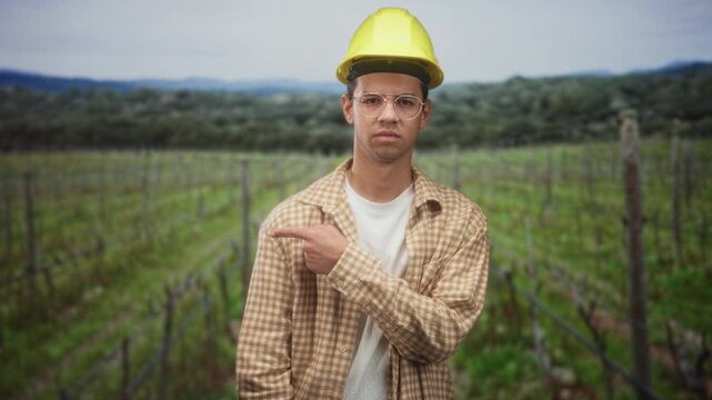 Man points finger to vines in vineyard wearing hardhat and glasses as construction worker; safety concern.