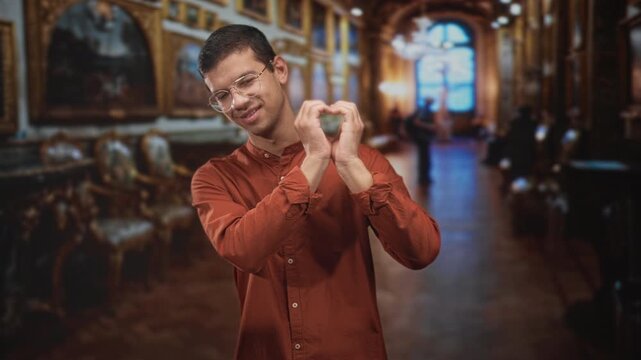 Man making a heart gesture with hands amid paintings and chairs in an ornate museum building; affection heritage.