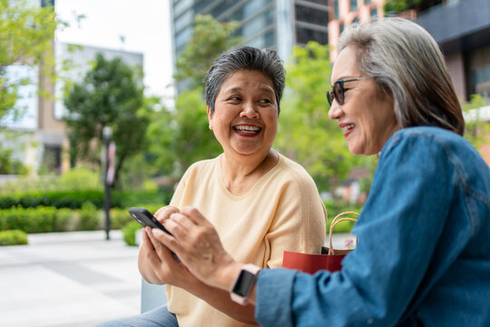 Two senior Asian women using phone shopping online outdoors in the city. 