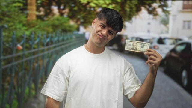 Young hispanic man holding us dollar bill between thumb and forefinger, pointing thumb to chest, smirking on street; confidence swagger.