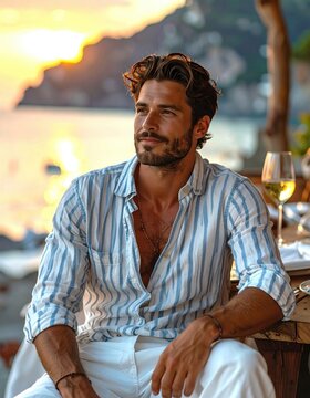 A relaxed man in a striped shirt enjoys a sunset moment by the water with a glass of white wine, surrounded by a scenic coastal backdrop.