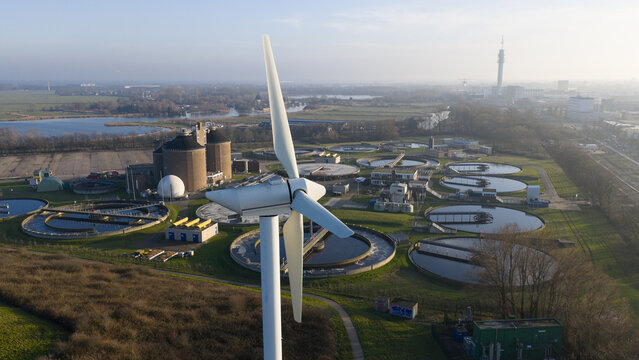 Aerial view of a large wind turbine standing near a water treatment facility with circular sedimentation tanks and industrial structures under a clear sky in Haarlem, Noord-Holland, Netherlands.