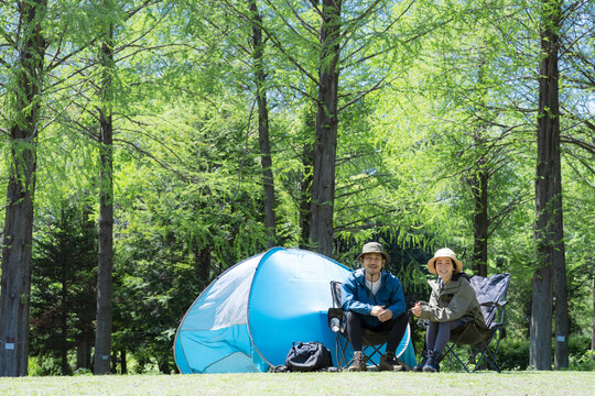 新緑のキャンプ場で椅子に座るカメラ目線笑顔のアウトドアやレジャーを楽しいミドルの夫婦全身の広角　左上にコピースペースあり