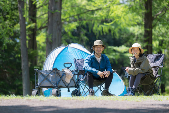 新緑のキャンプ場で椅子に座るカメラ目線笑顔のアウトドアやレジャーを楽しいミドルの夫婦全身