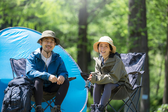 新緑のキャンプ場で椅子に座るカメラ目線笑顔のアウトドアやレジャーを楽しいミドルの夫婦