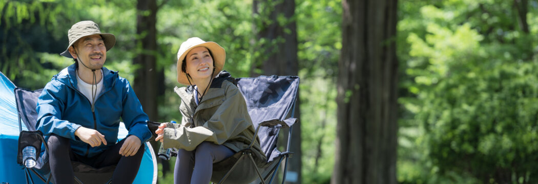 新緑のキャンプ場で椅子に座る笑顔のアウトドアやレジャーを楽しいミドルの夫婦　鳥や景色を眺めるバナー