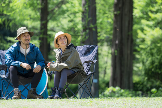 新緑のキャンプ場で椅子に座る笑顔のアウトドアやレジャーを楽しいミドルの夫婦　鳥や景色を眺める