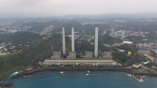 Aerial view of Concord Power Plant with its three tall chimneys situated on the rocky coastline against a backdrop of lush green hills in Xiehe Village, Keelung City, Taiwan.