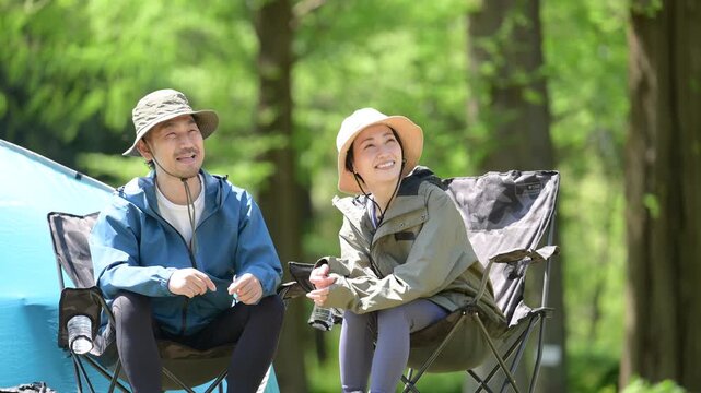 新緑のキャンプ場で椅子に座る笑顔のアウトドアやレジャーを楽しいミドルの夫婦　鳥や景色を眺める動画