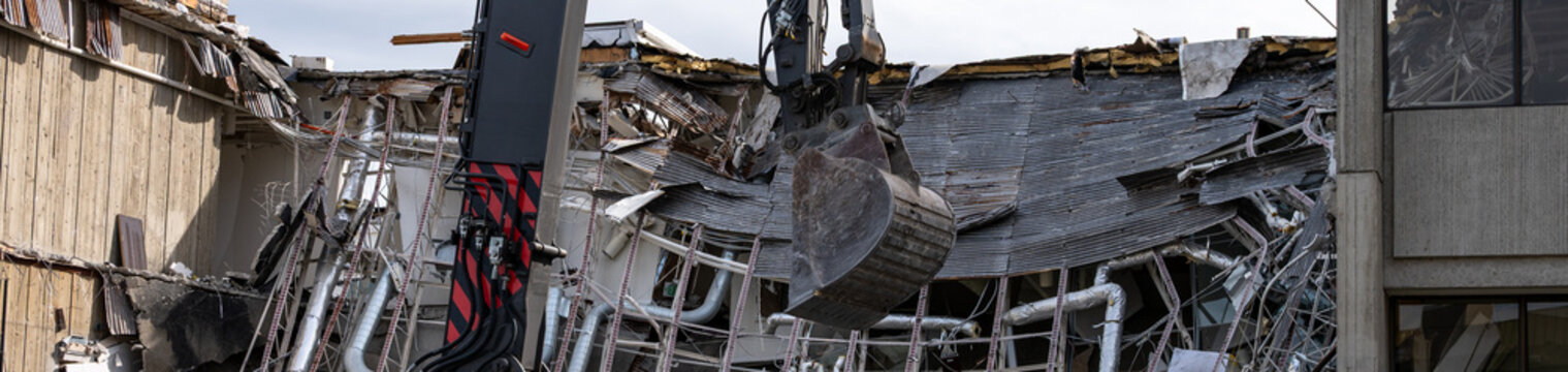 Heavy duty construction machinery to tear down old concrete office building for redevelopment, roof pulled down exposing heating and ventilation metal ducts and pipes
