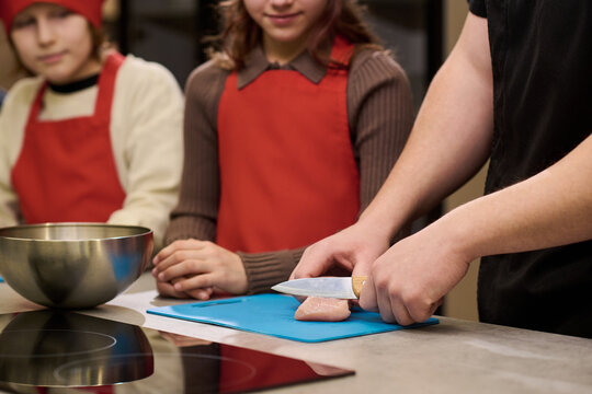 Caucasian adult chef slicing chicken on cutting board while children watching cooking lesson in kitchen. Suitable for culinary education, food safety training, parenting content