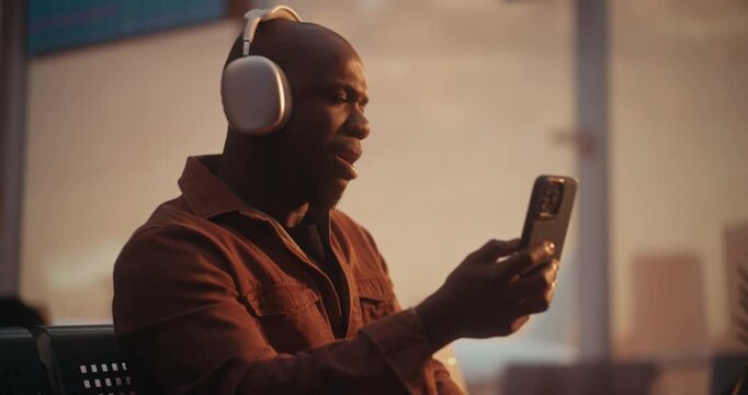 African American Male Traveler Conducts Virtual Meeting Via Mobile Device. With Headphones on, Man Focuses on Video Call, Nodding and Speaking Into Phone Microphone. Connected Traveler Lifestyle.