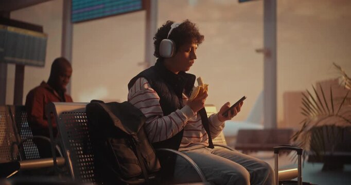 Young Man With Curly Hair Enjoys Healthy Snack While Waiting For Flight. Male Sits in Airport Terminal, Wearing Large Silver Over Ear Headphones, Taking Bite of Banana While Focused on Smartphone.