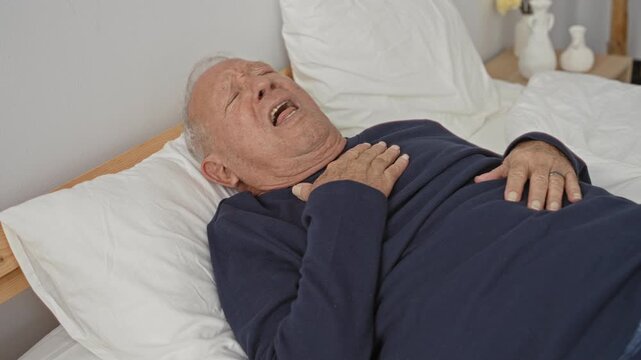 Old man clutching chest on bed in a bedroom building, mouth open and hand pressing sternum; pain emergency.