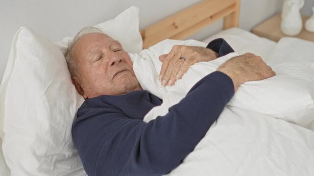 Man lying in bed holding duvet with hand on chest, grey haired senior sleeping, wedding ring visible, wooden headboard and white sheets in a bedroom inside a building; serenity.