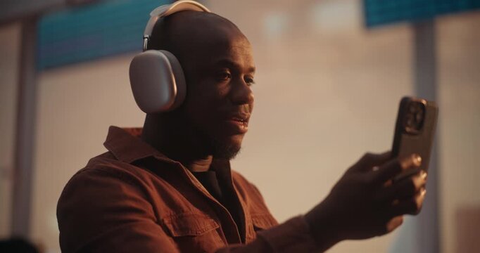 Black Man, Wearing Stylish Silver Over Ear Headphones and Overshirt, Engages in Video Chat on Smartphone. Sitting in Airport Departure Lounge, Man Smiles and Sharing Travel Excitement With Loved Ones.