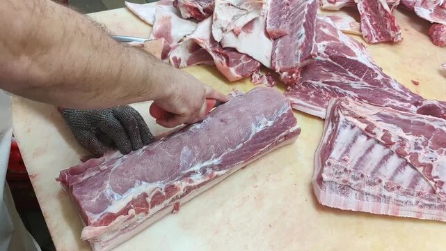 Butcher prepares pork cuts on a wooden table, skillfully slicing through meat with a knife, showcasing various pork pieces and tools in a clean workspace