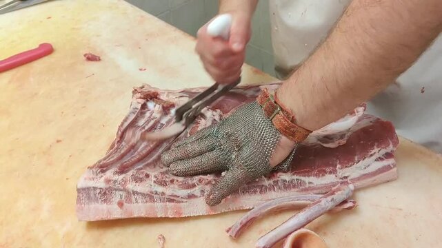 Male butcher in protective gloves skillfully cuts pork belly on a wooden countertop, showcasing precise knife techniques and meat preparation in a clean kitchen environment