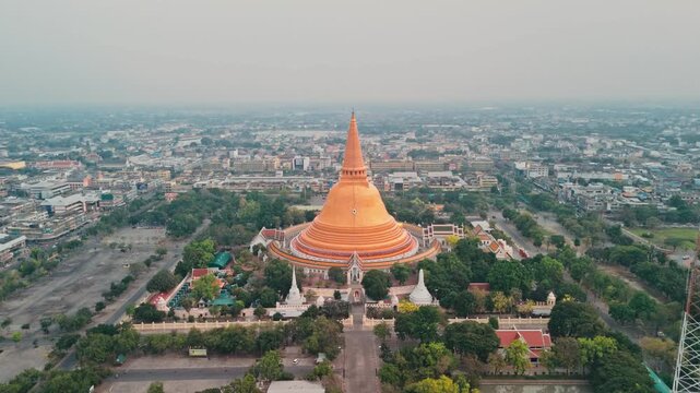 High angle drone perspective pulls away from huge golden stupa rising above urban center while majestic Buddhist monument dominates hazy gray city skyline in Thailand during serene beautiful morning