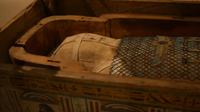 A mummy resting inside an ancient Egyptian sarcophagus adorned with detailed hieroglyphics and ornate carvings. Close-up view highlighting aged textures, preserved remains