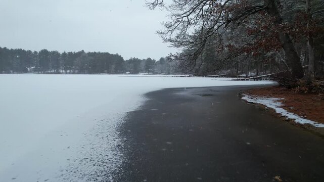 Low altitude drone flight over icy edge of frozen lake during snowfall in Harold Parker State Forest, Massachusetts, USA. Aerial forward