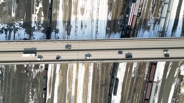 Top down drone shot of a snowy bridge highway above sprawling train tracks as the camera lifts and cars move below