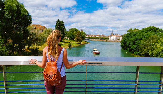 Woman traveler with leather backpack looking at Saone river and boat from a bridge in Chalon-sur-Saone, Burgundy, France. Summer travel lifestyle and river tourism in Saone-et-Loire.